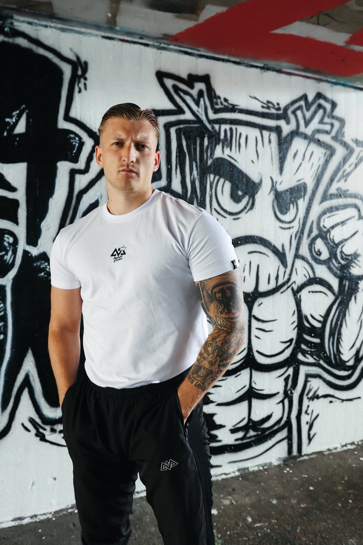 Man wearing white Avantura T-shirt with small chest logo against graffiti wall