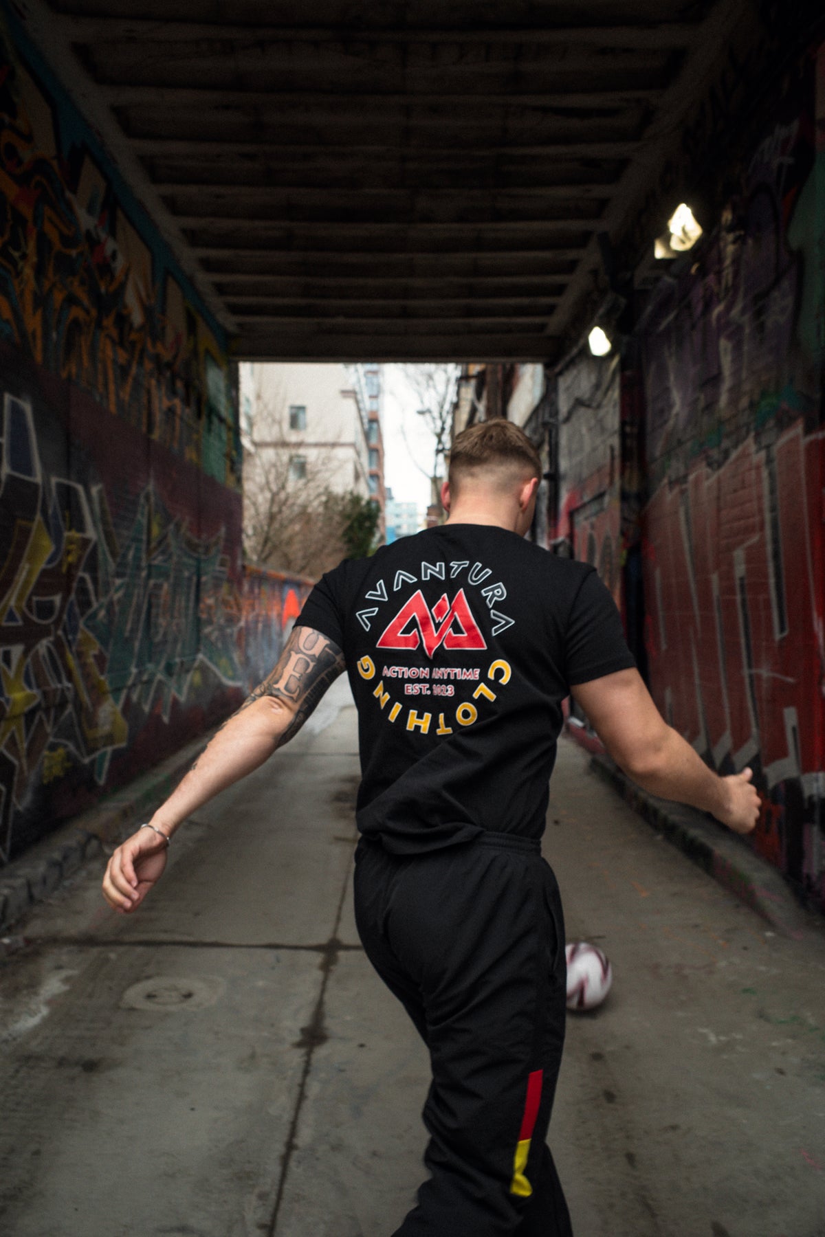 Man walking away in black Avantura T-shirt with large circular back graphic, soccer ball visible in urban tunnel setting