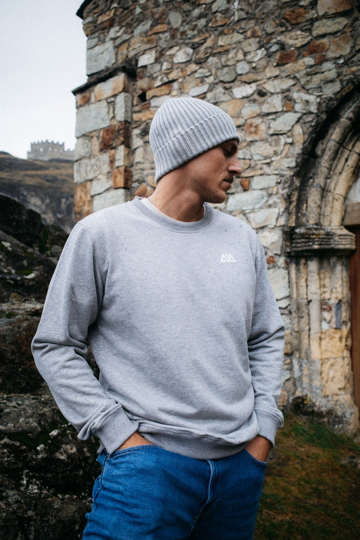 Mann in grauem Crewneck Sweater mit Logo und Mütze vor Steinruine