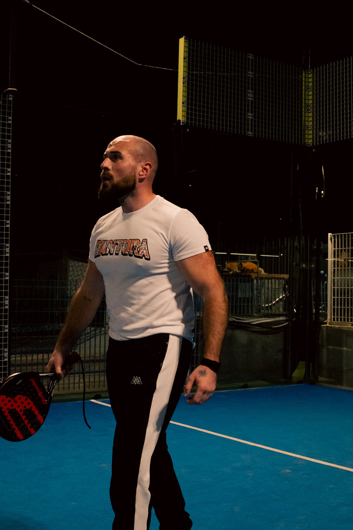 Bearded man on padel court in white Avtura T-shirt with multicolor trim