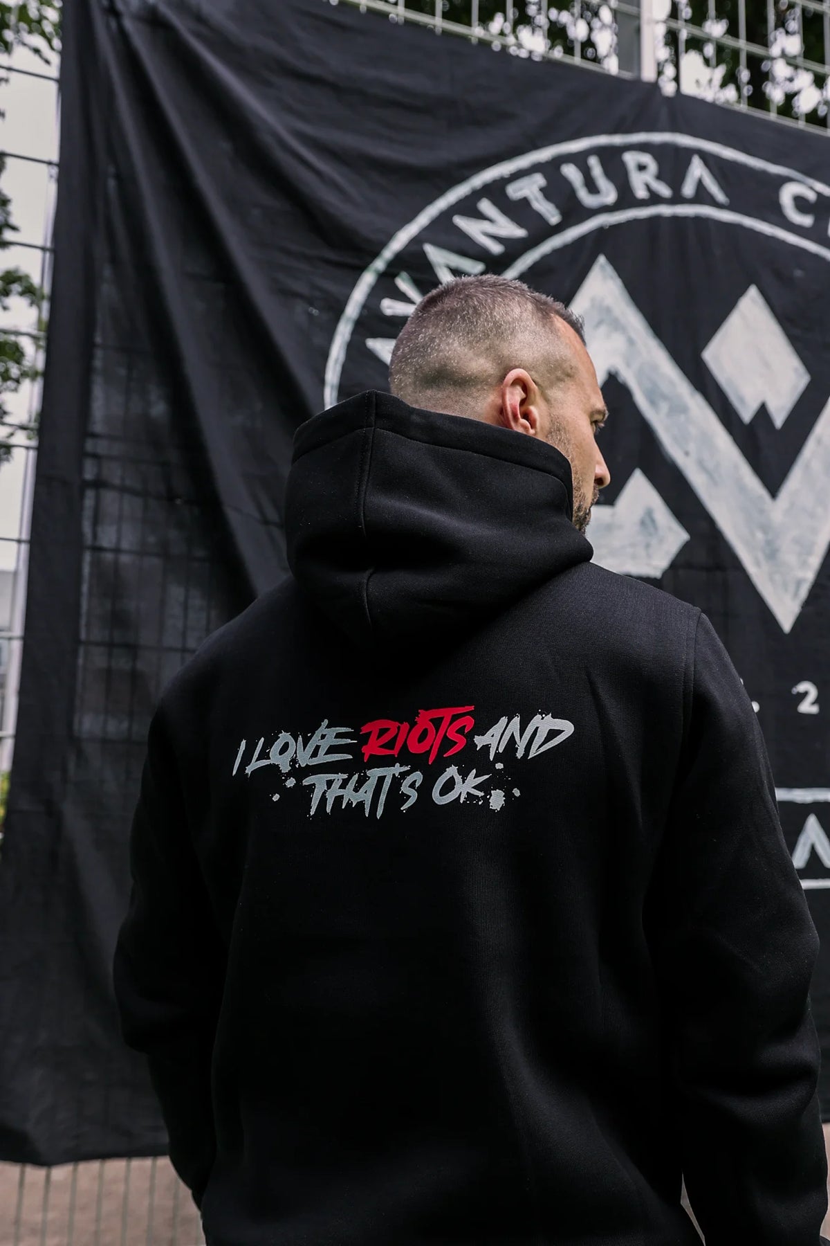 Side view of man in black hoodie showing hood down and large back 'I LOVE RIOTS' print