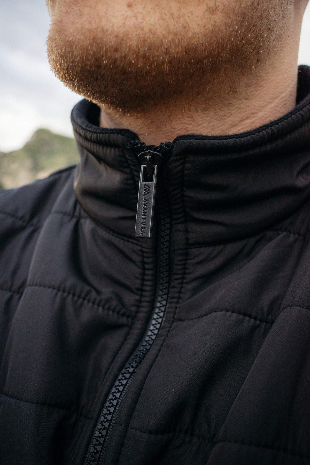 Close-up of black fleece jacket zipper with metal AVANTURA pull tab and high collar