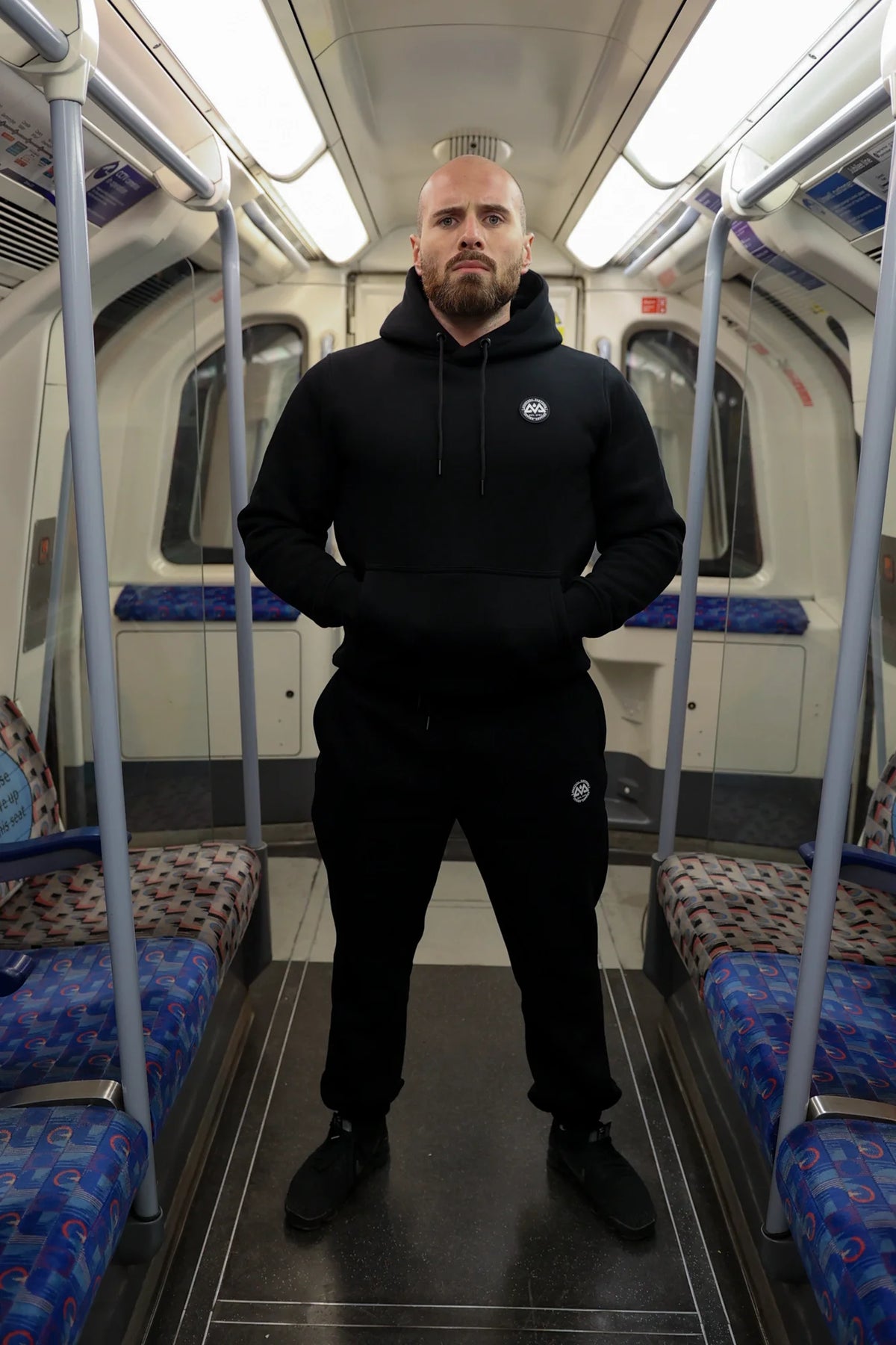 Man wearing black hoodie and jogger pants set in subway, hands in front pouch pocket