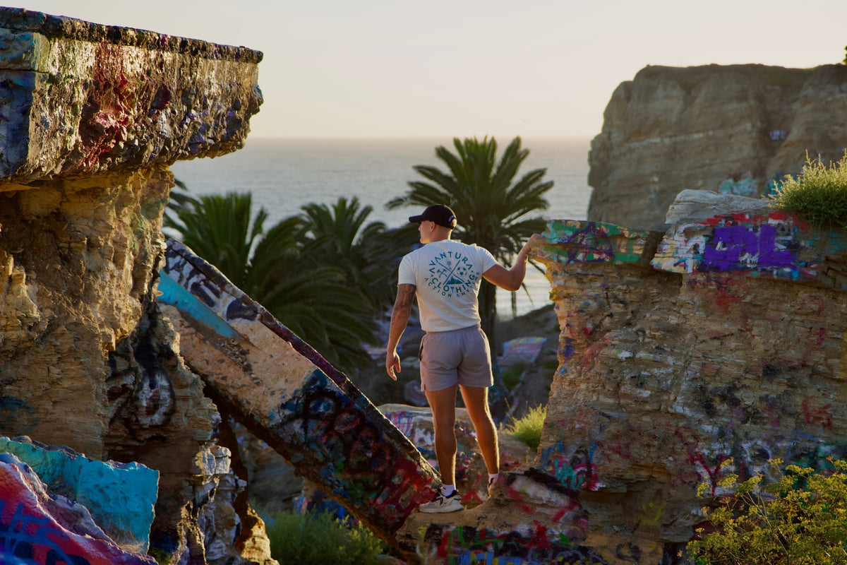 Beige Avantura lifestyle t-shirt with circular back logo, worn by man on coastal ruins