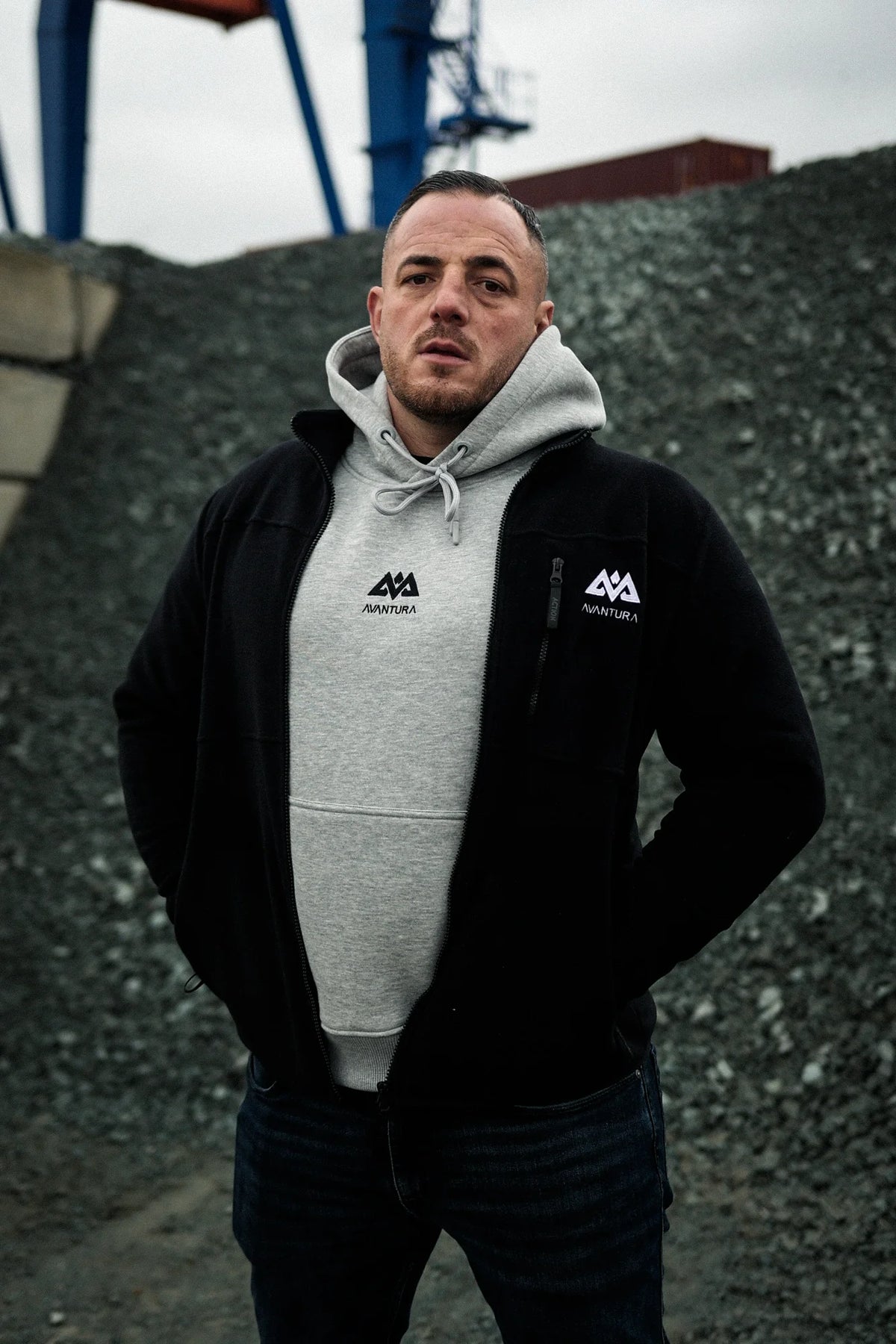 Front view of man wearing heather grey hoodie under open black jacket showing chest logo