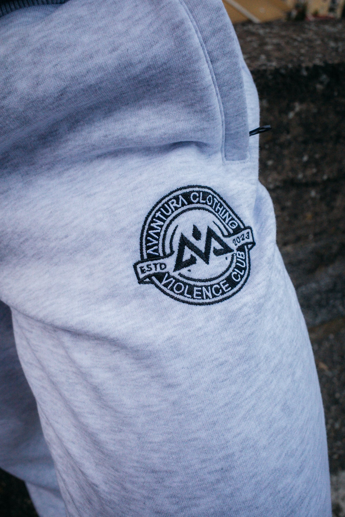 Close-up of black embroidered AVANTURA CLOTHING logo on light grey heathered jogger fabric