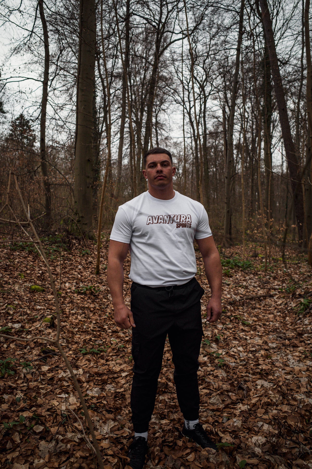 Full body shot of man modeling white Avantura T-shirt with chest logo in forest