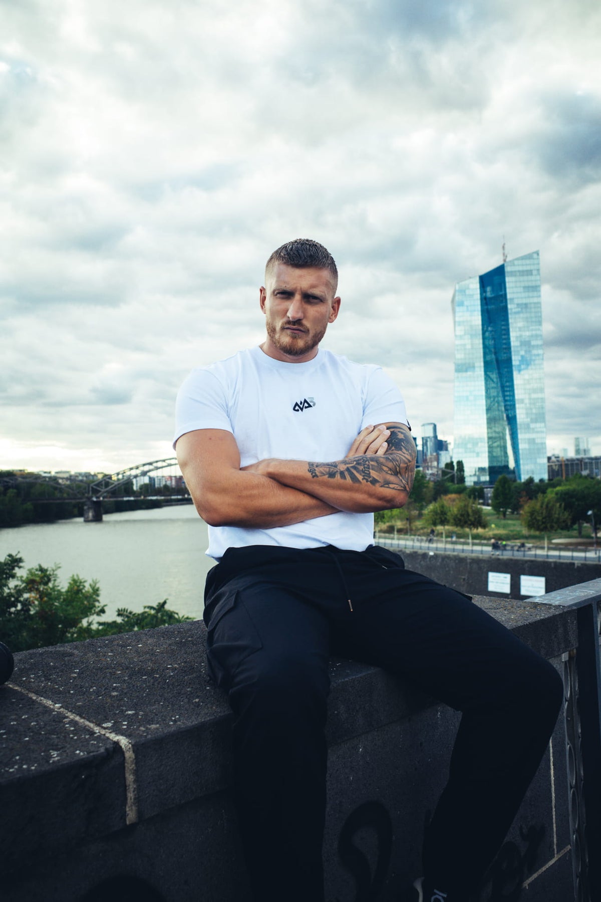 Weißes Avantura T-Shirt mit kleinem Brustlogo, getragen von Mann in Stadtlandschaft