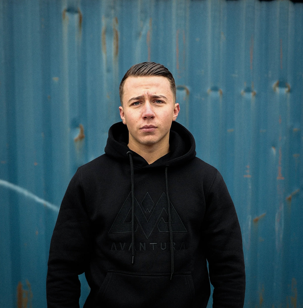Schwarzer AVANTURA Hoodie mit gesticktem Logo, getragen von einem Mann vor blauer Wellblechwand