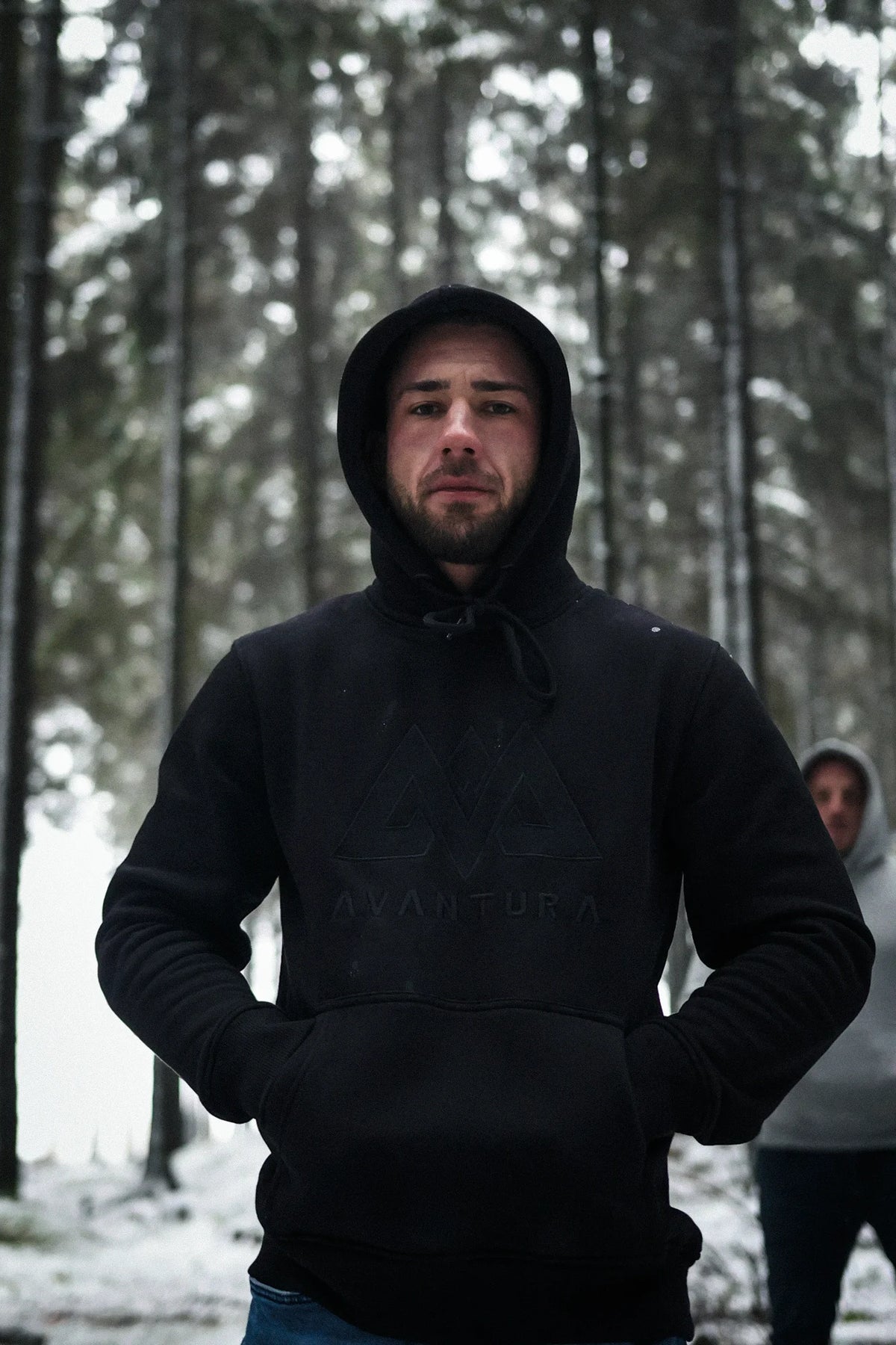 Schwarzer AVANTURA Hoodie mit Kapuze in schneebedecktem Wald, Hände in der Tasche