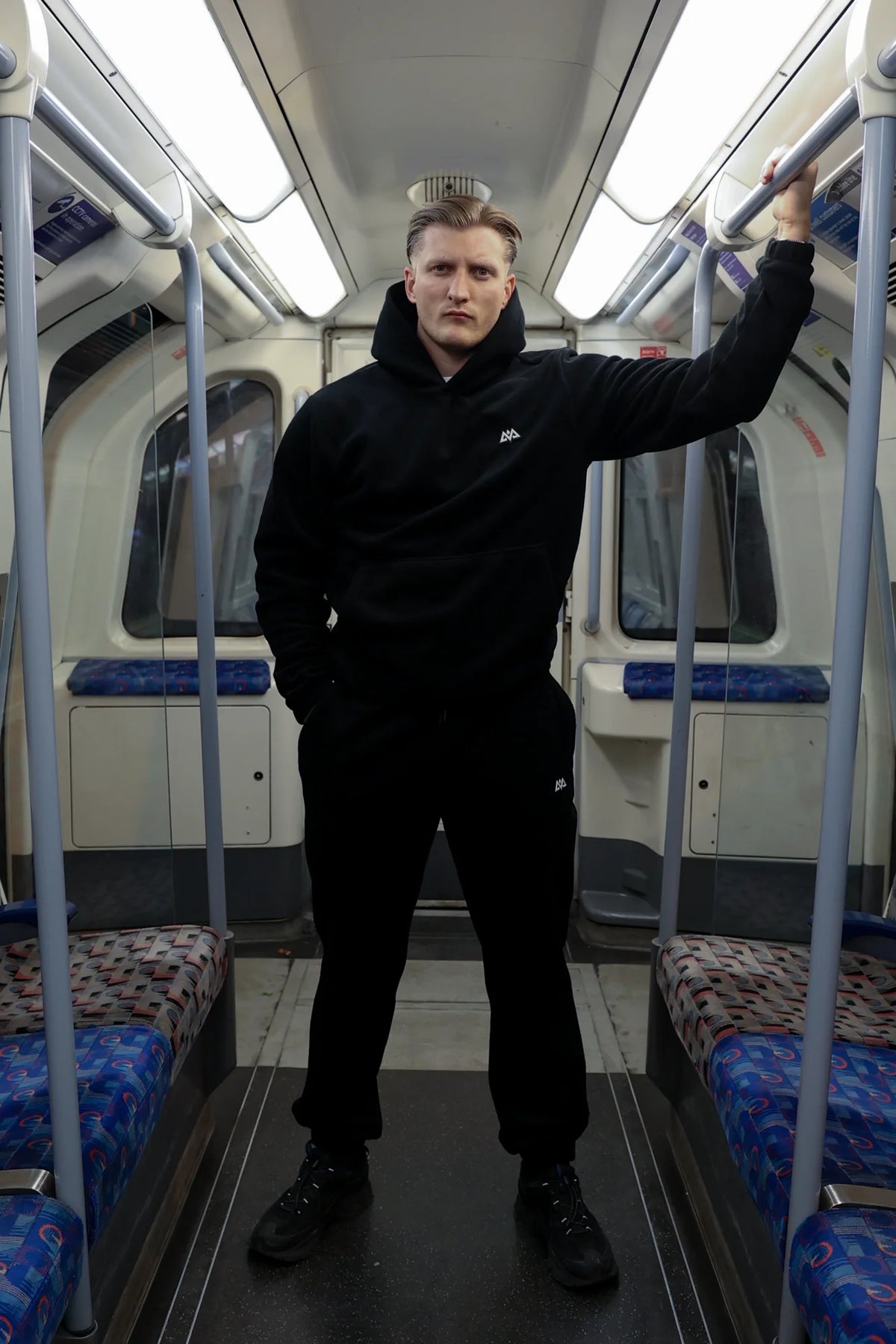Full body shot of black fleece hoodie outfit with white logo holding subway bar