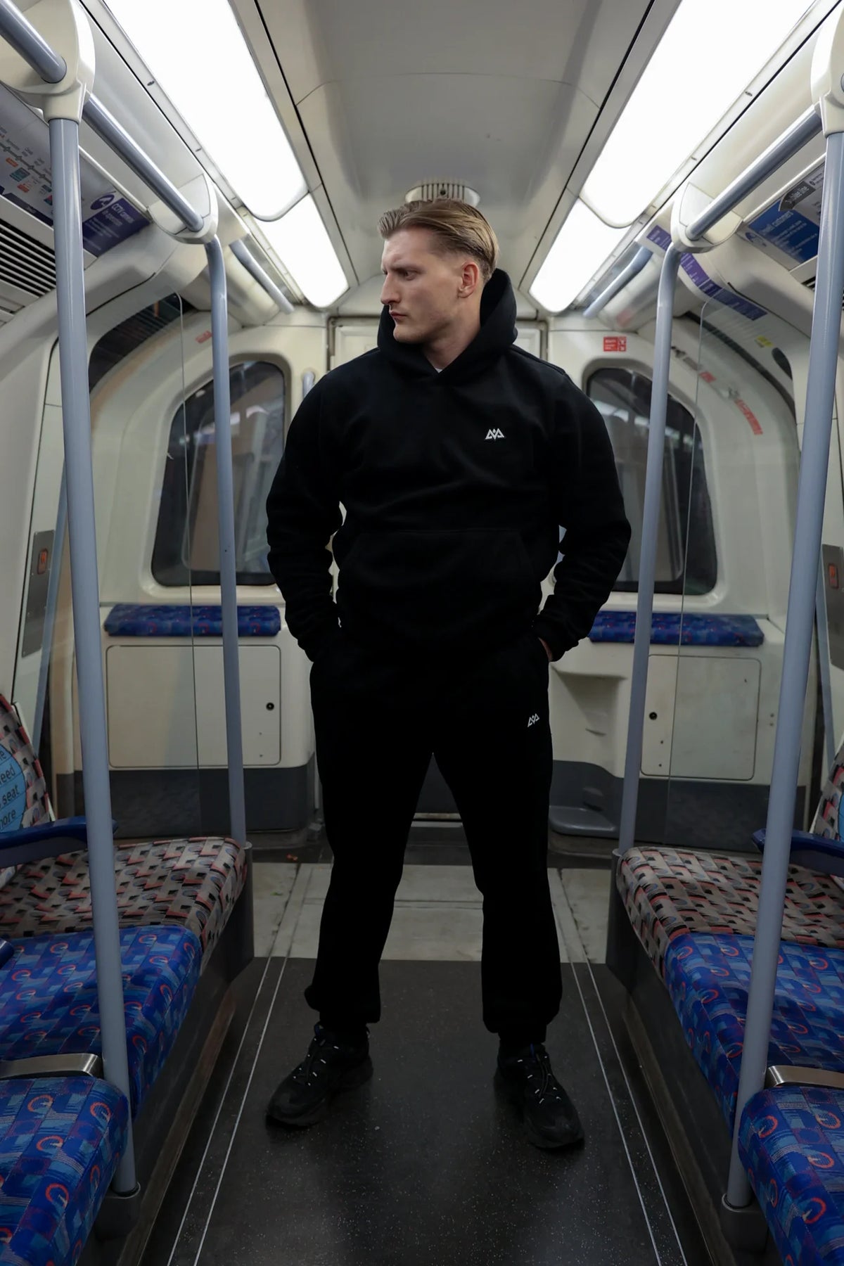 Man wearing black AVA Logo Fleece Hoodie and sweatpants standing in subway carriage