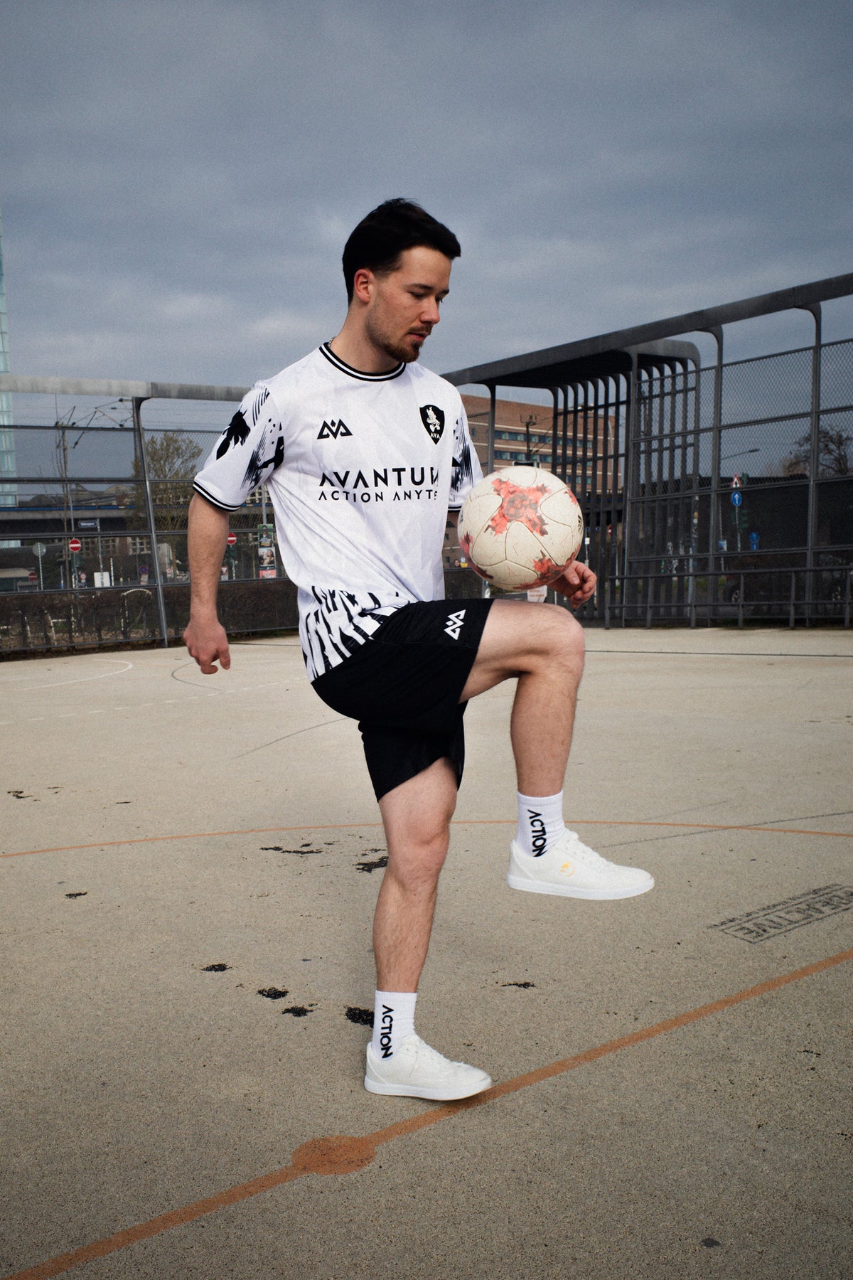 Front view of man in white Avantura jersey and black shorts holding soccer ball on urban court