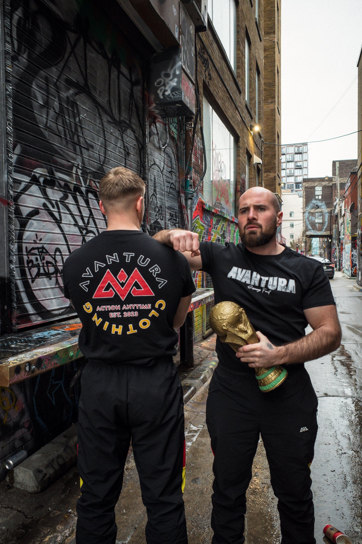 Man holding FIFA trophy wearing black Avantura T-shirt with stylized chest print, standing in graffiti alley with partner