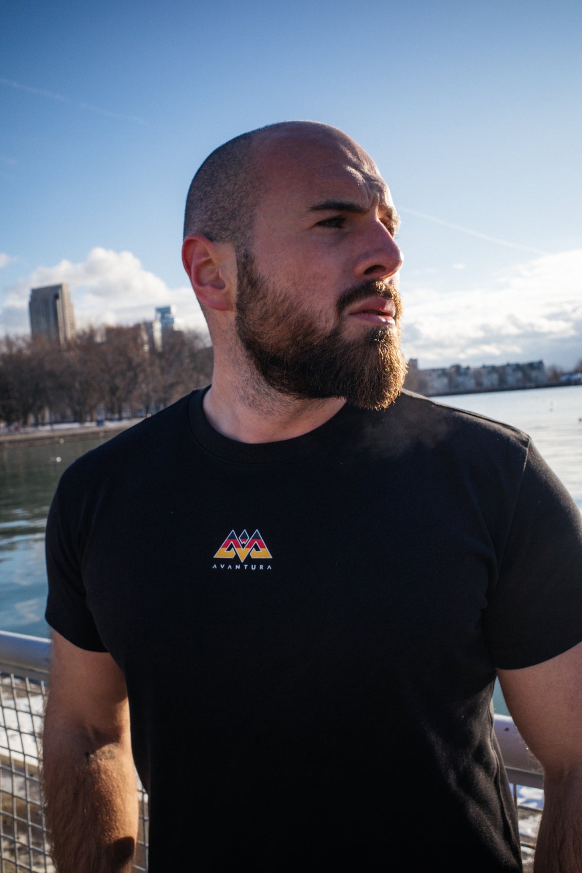 Close-up of black crew-neck T-shirt showing small colorful geometric AVANTURA logo on left chest area