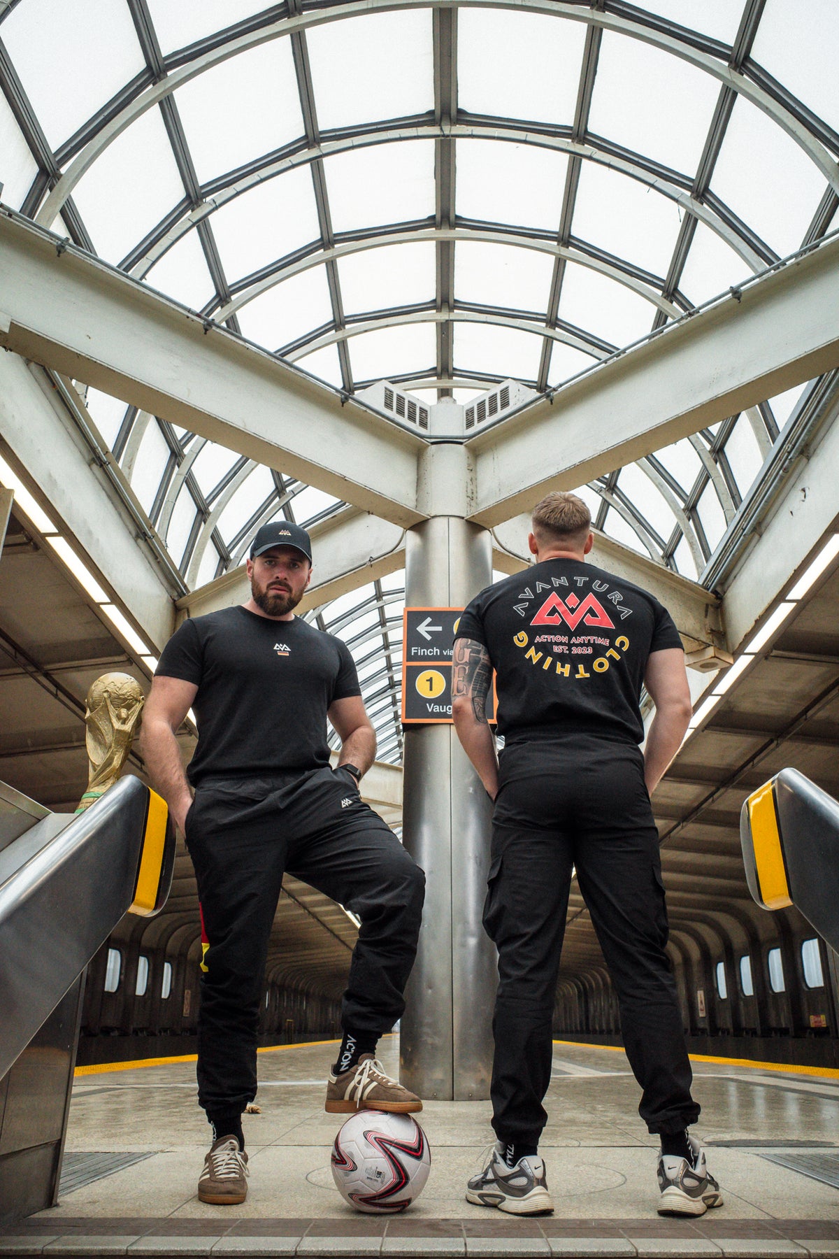 Two men in subway station wearing black Avantura T-shirts with front chest logo and large circular back print design