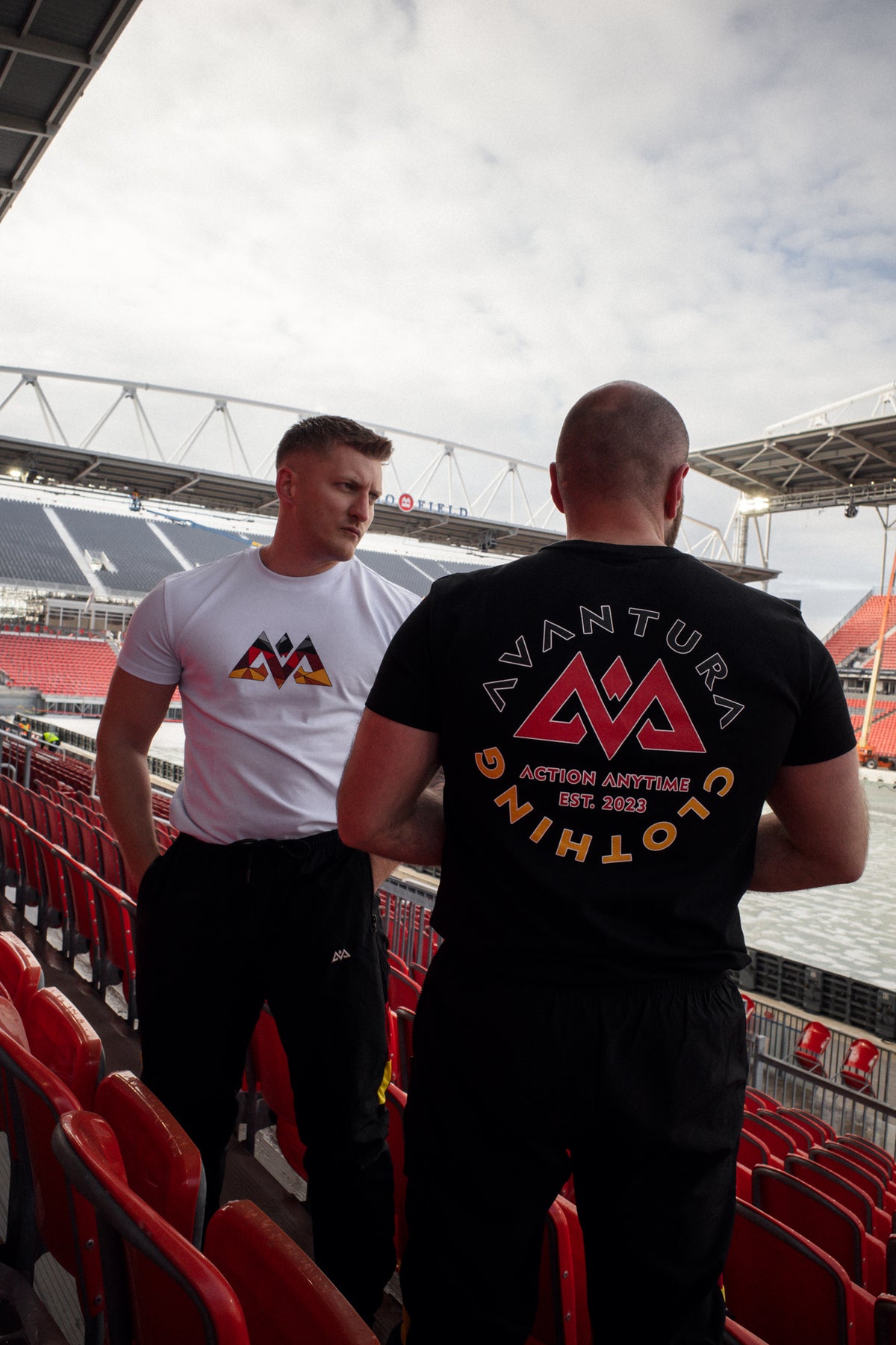 Men wearing black and white Avantura T-shirts in stadium seating, showcasing front chest logo and large back graphic design