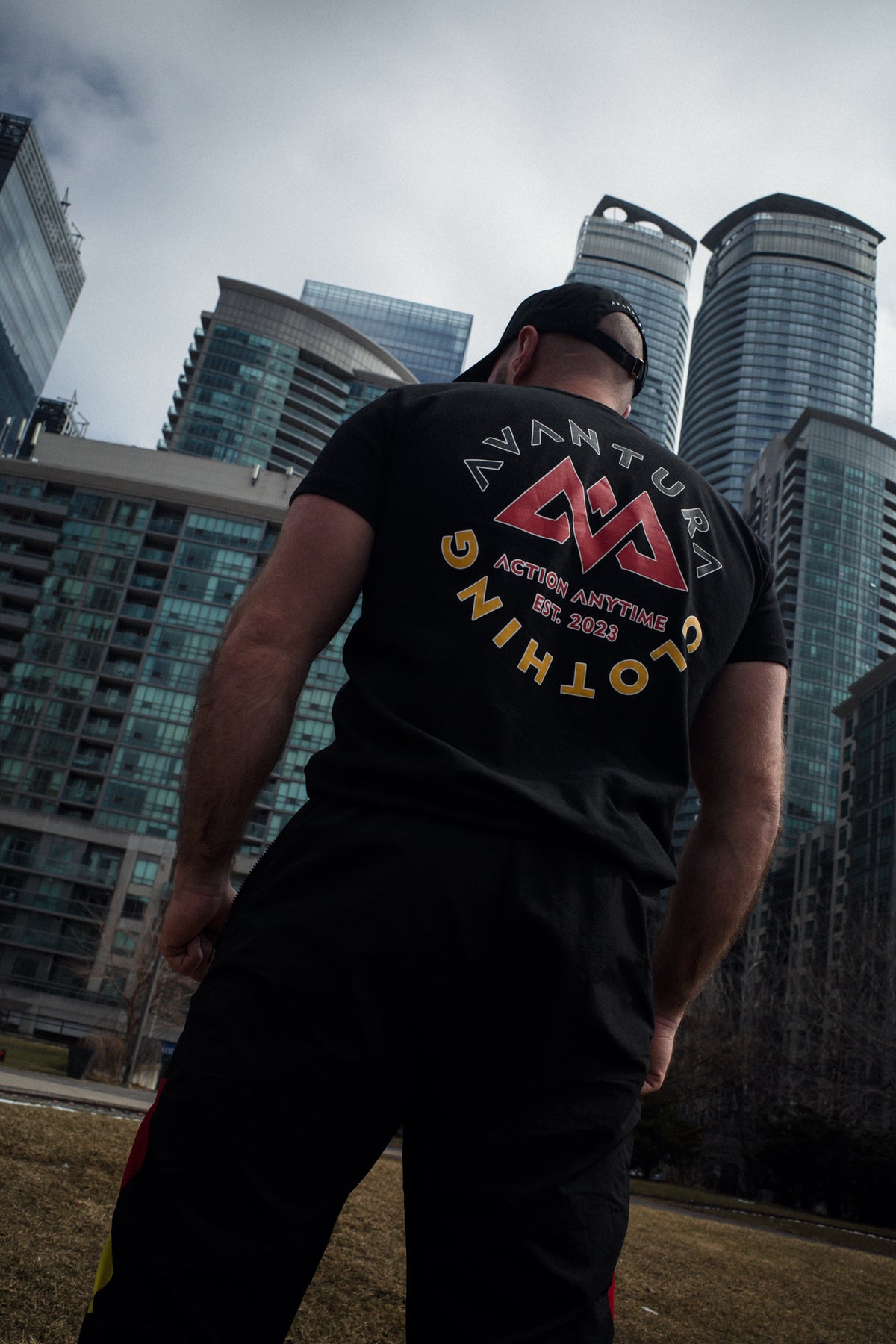 Back view of black Avantura T-shirt with large circular 'Action Anytime' back print against urban glass building backdrop