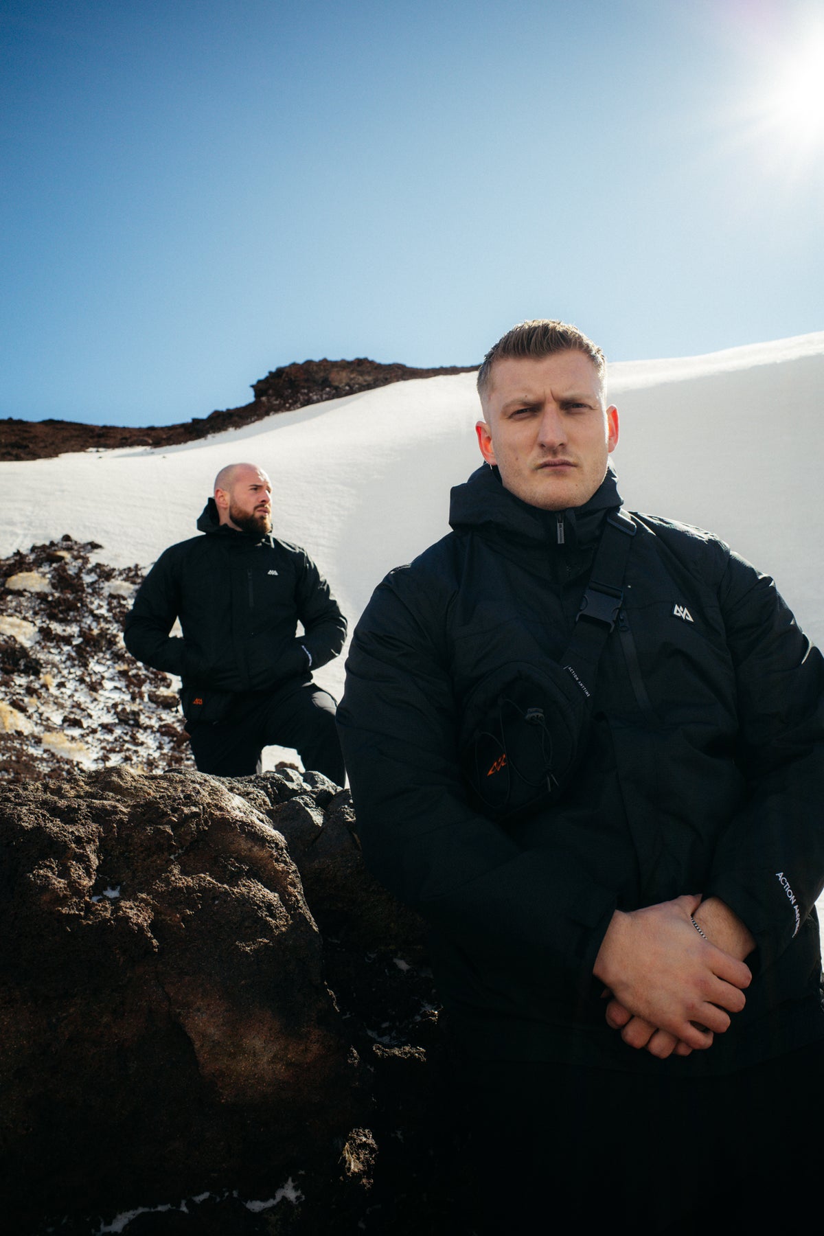 Two men modeling black technical jackets with mountain logo in sunlight