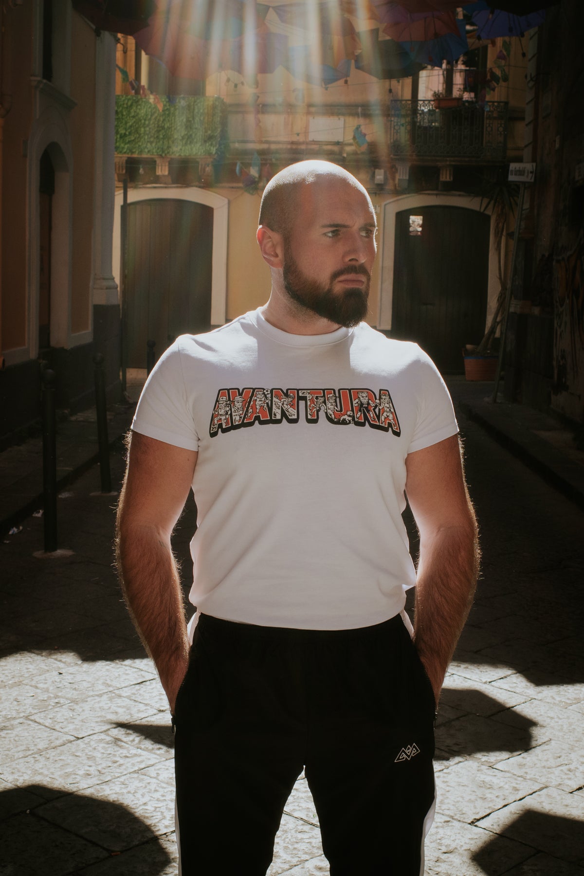 Mann trägt weißes T-Shirt mit buntem Avventura-Logo in sonniger Gasse