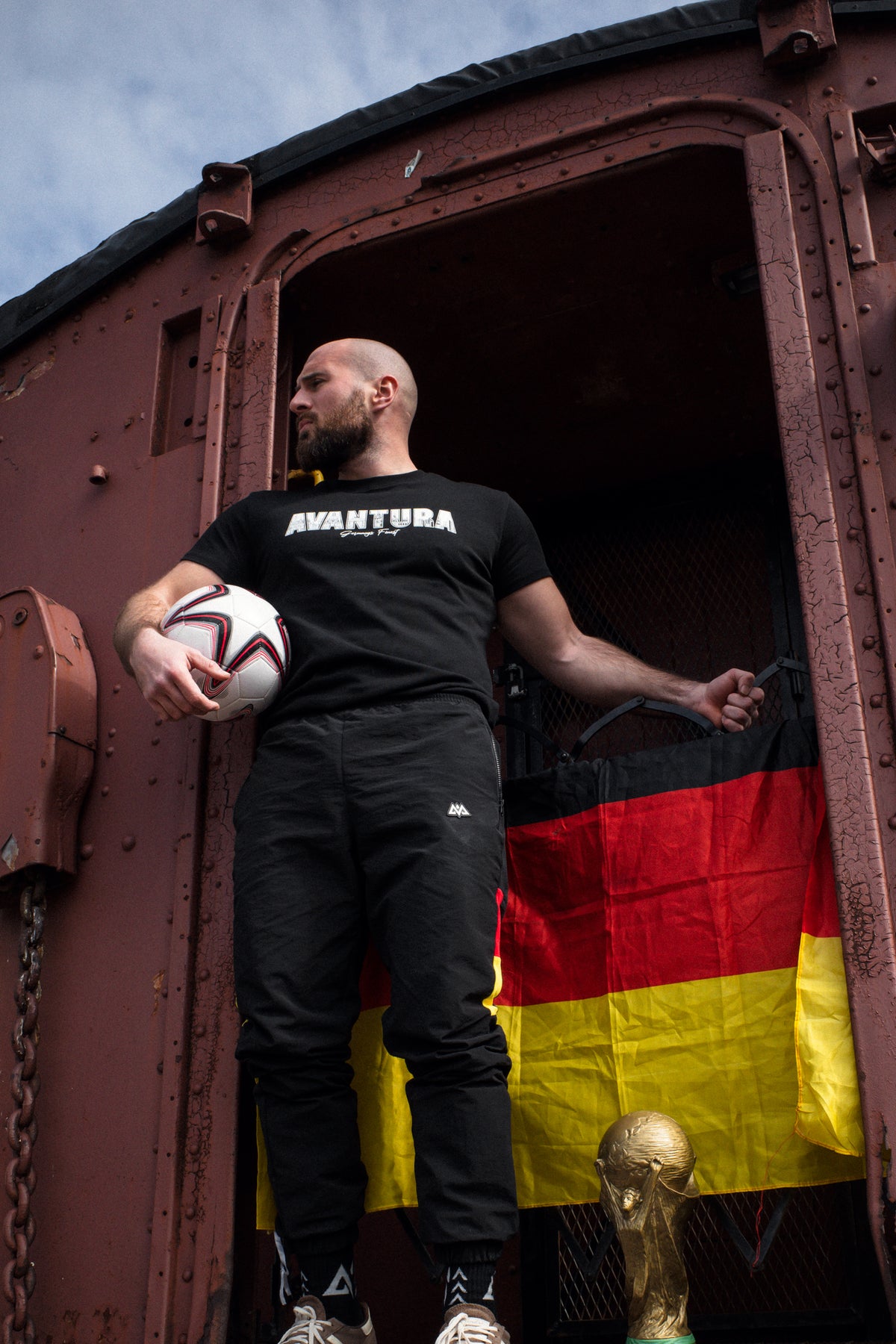 Avantura T-Shirt Schwarz getragen von unten mit WM-Trophäe im Hintergrund