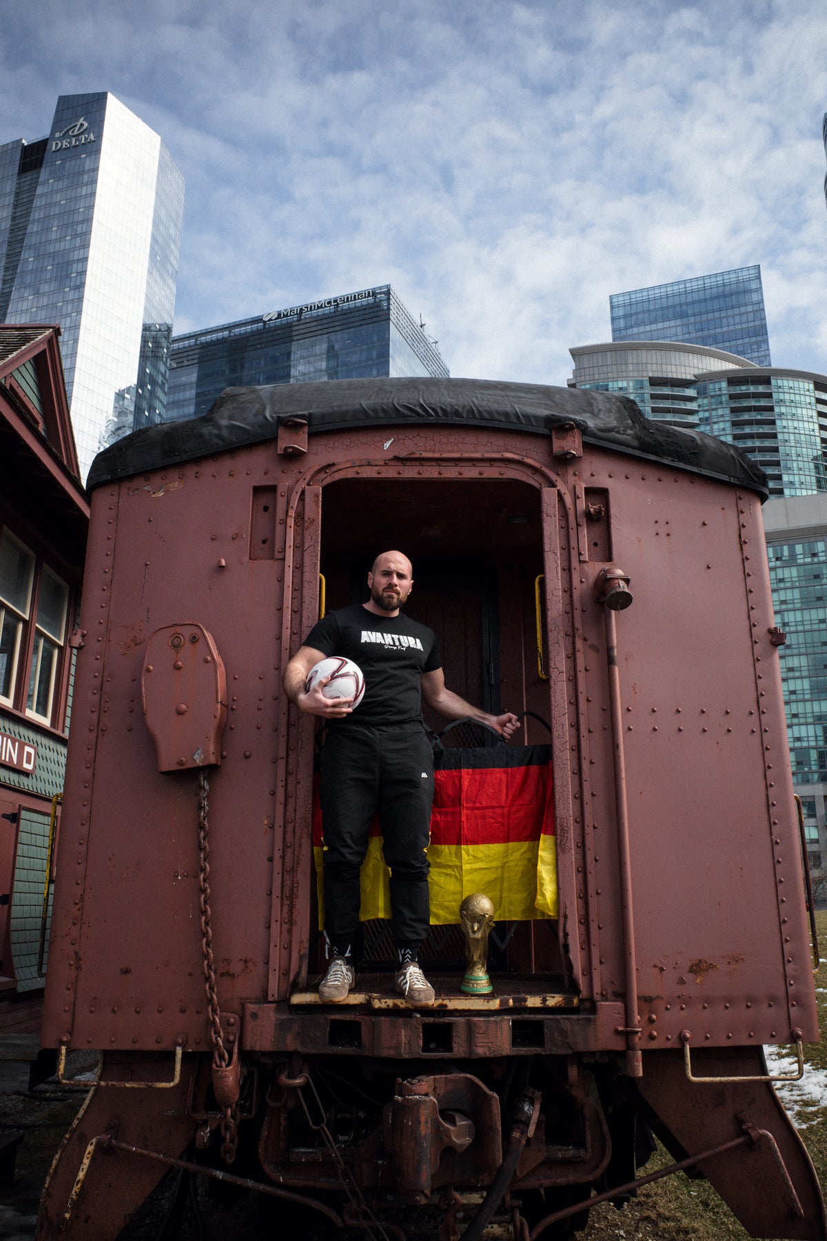 Mann in Avantura T-Shirt Schwarz mit Fußball vor Eisenbahnwagen