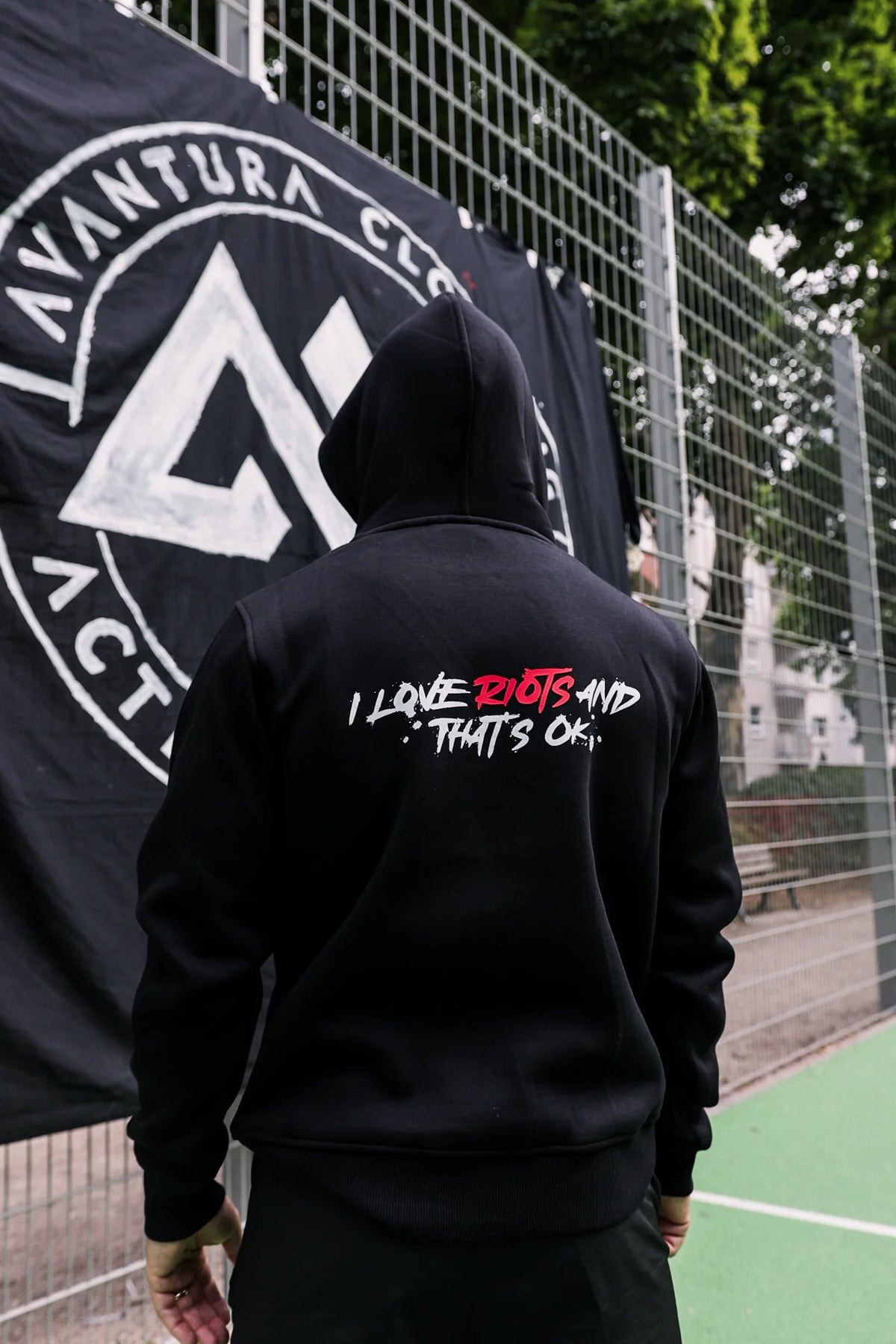 Person wearing black hoodie with hood up, displaying full back graphic and wire fence background