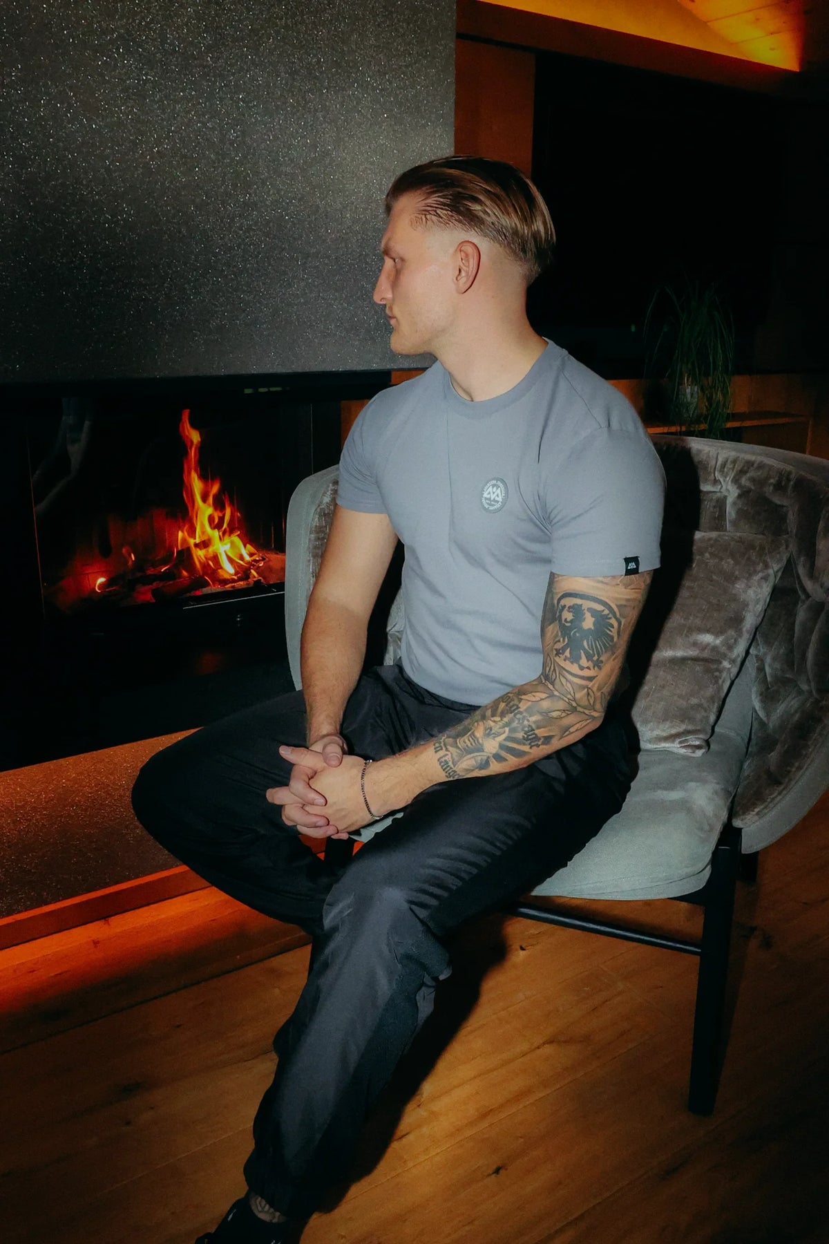 Grey crewneck T-shirt with chest logo, worn by a man in front of a fireplace