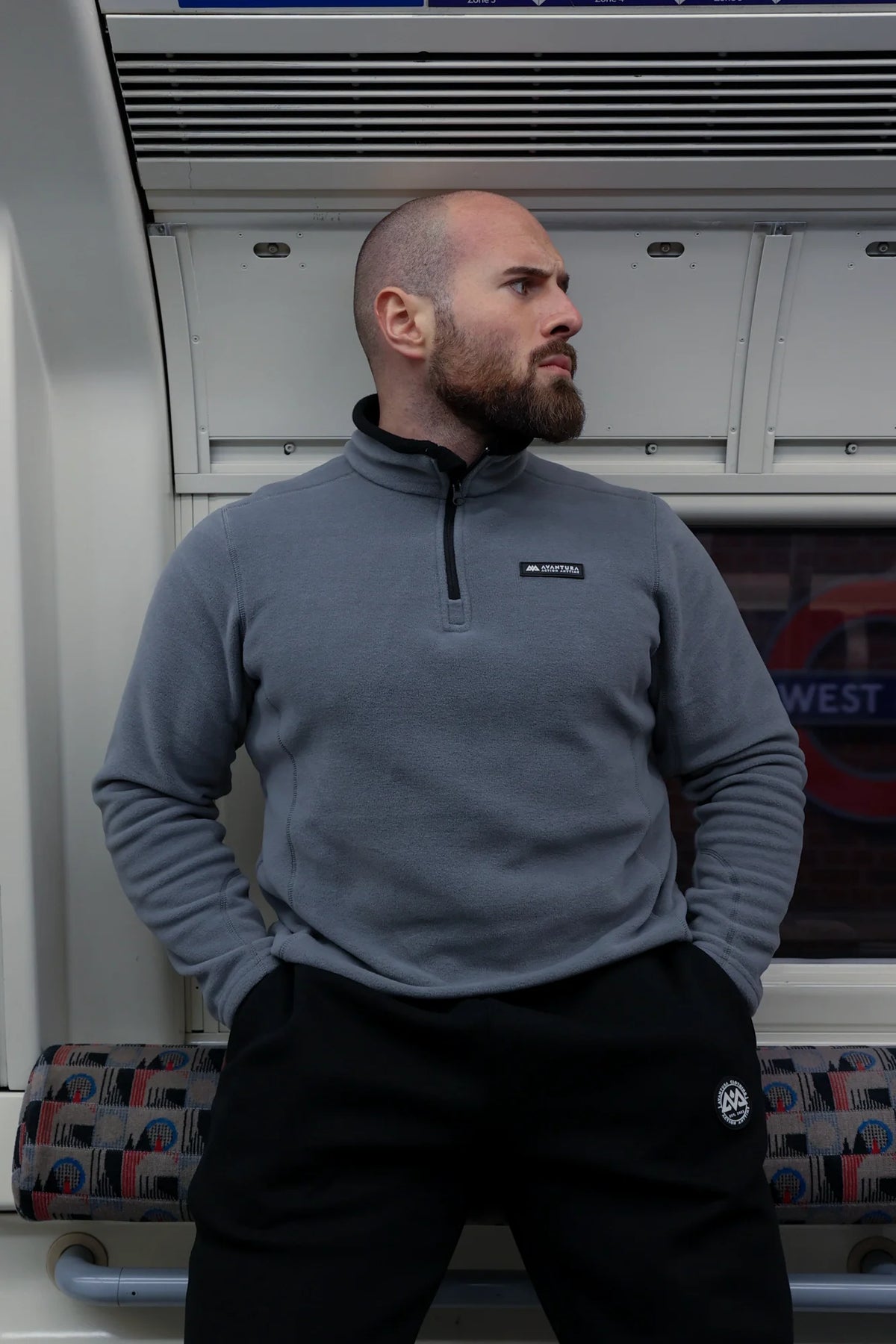 Man wearing grey halfzip fleece sweater in train, showcasing casual fit and black chest logo