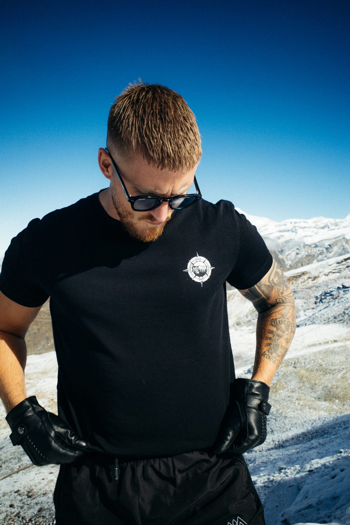 Black T-shirt with small white chest logo against snowy mountain backdrop