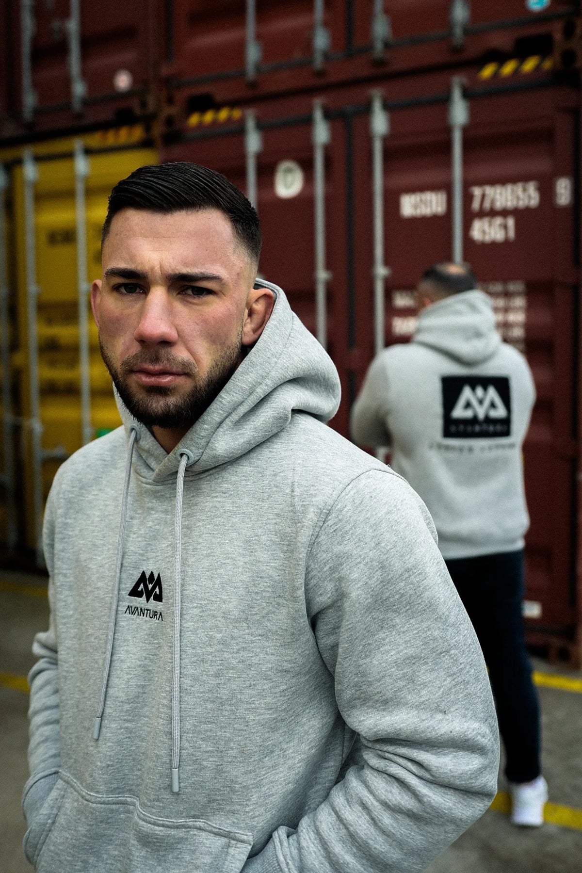 Two men in heather grey hoodies showing front logo and large back print in industrial setting