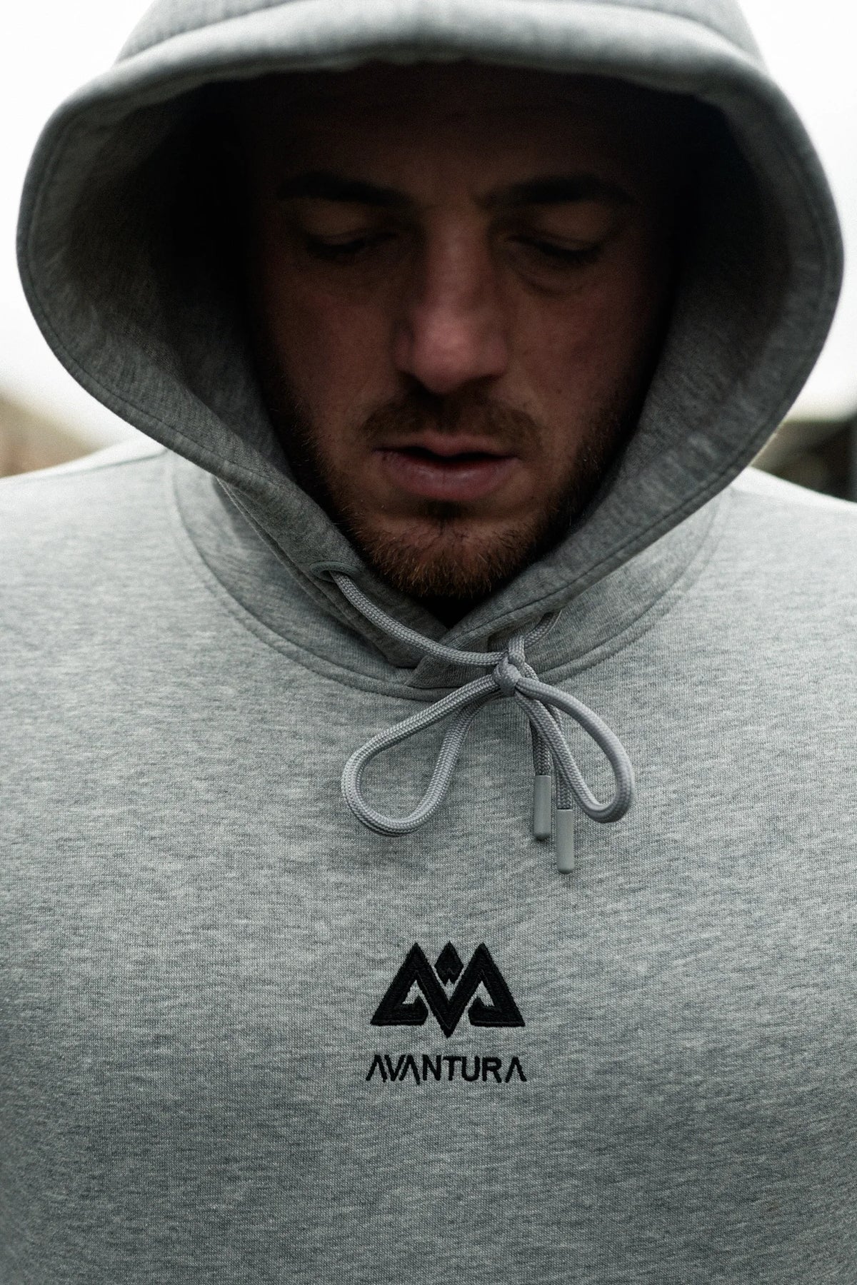 Man wearing heather grey hoodie with hood up, showing centered chest logo detail