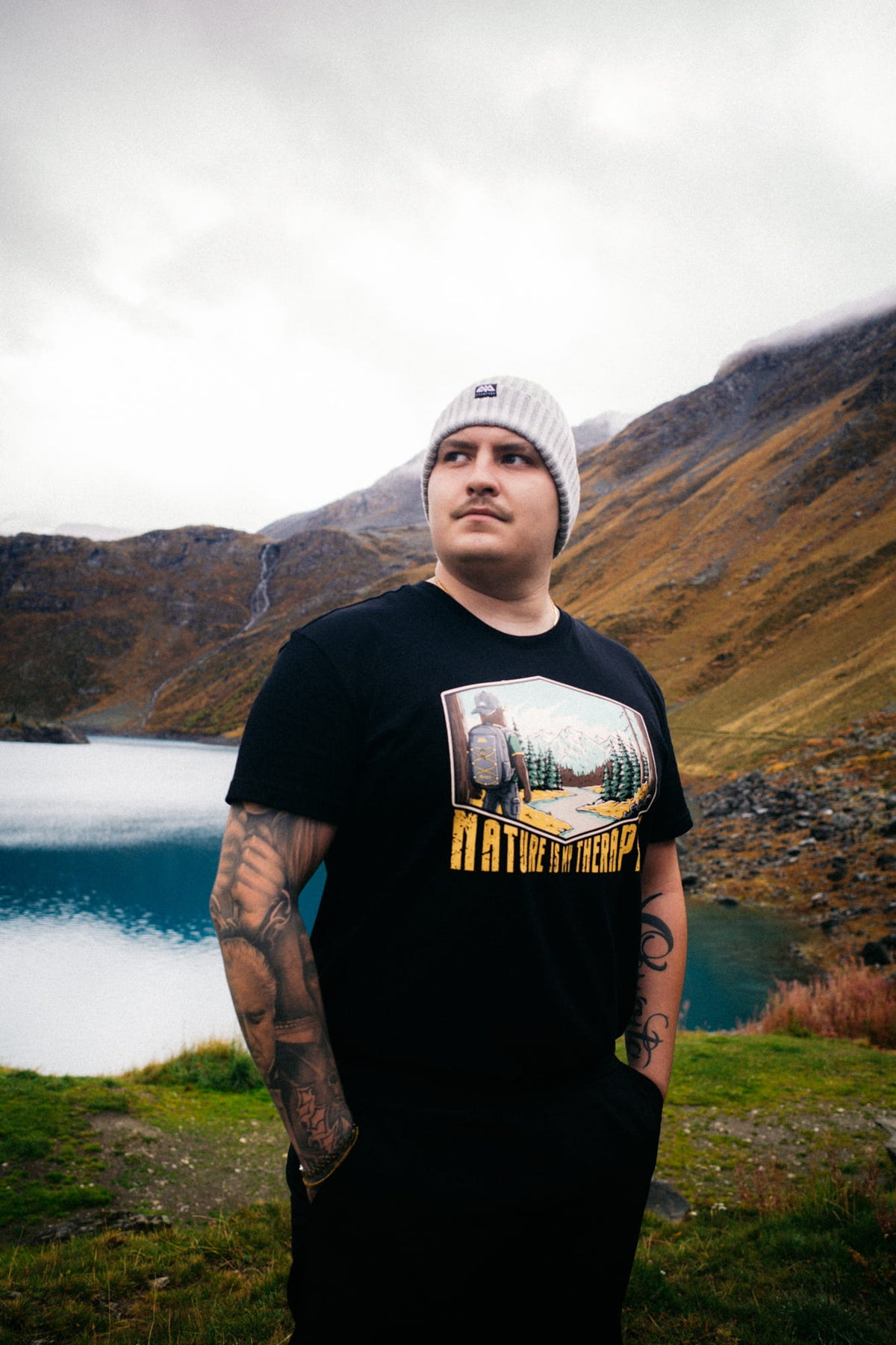 Tattoierter Mann in schwarzem T-Shirt mit Rucksack-Illustration am See